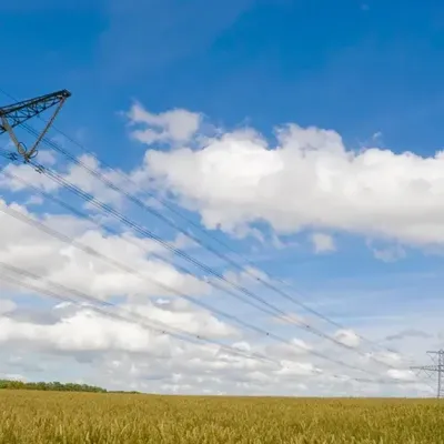 High-voltage power lines run across a wheat field under a blue sky with scattered clouds. The metal towers stretch into the distance, and the golden wheat covers the foreground.